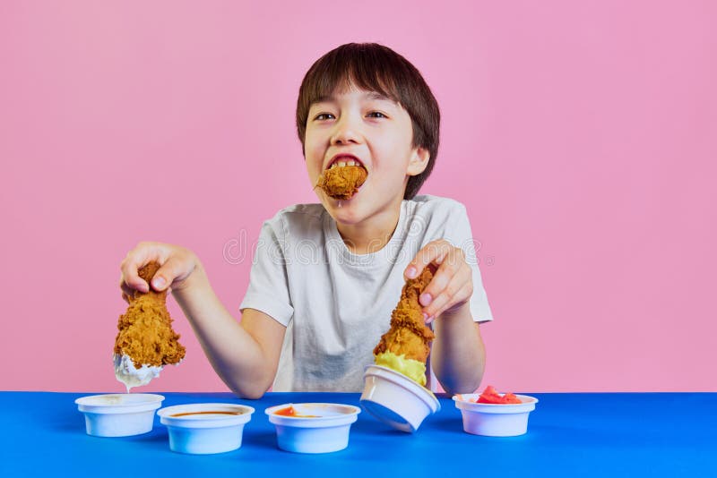 Little boy, child in white t-shirt sitting at table, dipping fried chicken in mustard and mayonnaise sauce and emotionally eating against pink background. Concept of food, childhood, emotions, pop art. Little boy, child in white t-shirt sitting at table, dipping fried chicken in mustard and mayonnaise sauce and emotionally eating against pink background. Concept of food, childhood, emotions, pop art