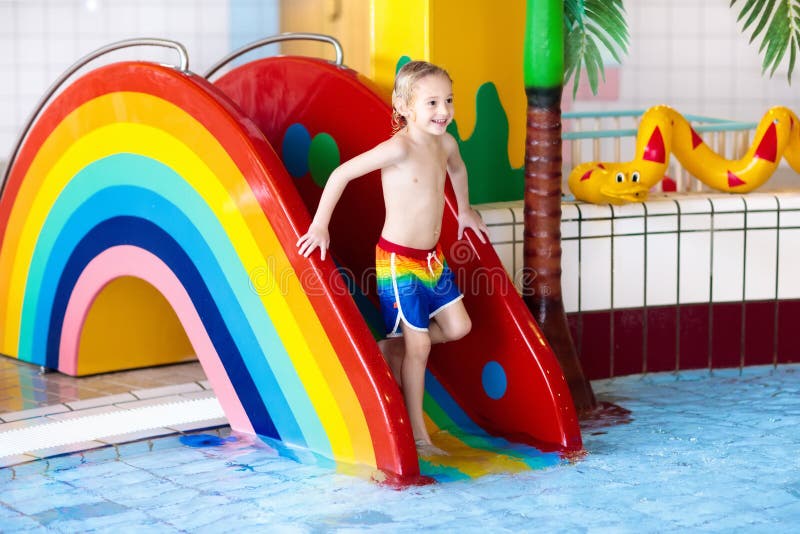 Child on swimming pool slide. Kid having fun sliding in water amusement park. Kids swim. Family summer vacation in tropical resort. Little boy in baby pool with colorful rainbow water slide. Child on swimming pool slide. Kid having fun sliding in water amusement park. Kids swim. Family summer vacation in tropical resort. Little boy in baby pool with colorful rainbow water slide.