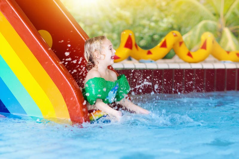 Child on swimming pool slide. Kid having fun sliding in water amusement park. Kids swim. Family summer vacation in tropical resort. Little boy in baby pool with colorful rainbow water slide. Child on swimming pool slide. Kid having fun sliding in water amusement park. Kids swim. Family summer vacation in tropical resort. Little boy in baby pool with colorful rainbow water slide.