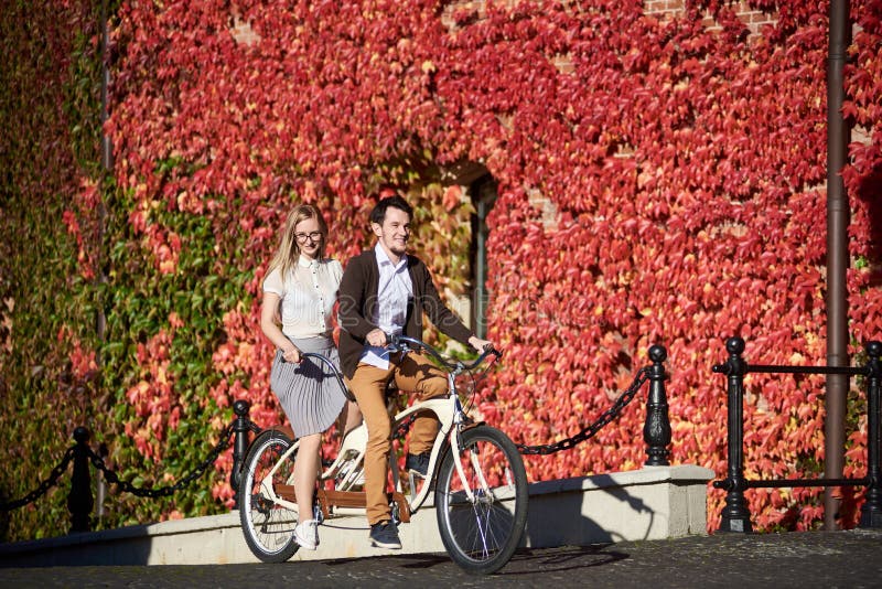 Barnet kopplar ihop, den stiliga mannen och blonda kvinnan som cyklar den tandema cykeln, genom att bygga som är bevuxet med den fotografering för bildbyråer Barnet kopplar ihop, den stiliga mannen och blonda kvinnan som cyklar den tandema cykeln, genom att bygga som är bevuxet med den fotografering för bildbyråer