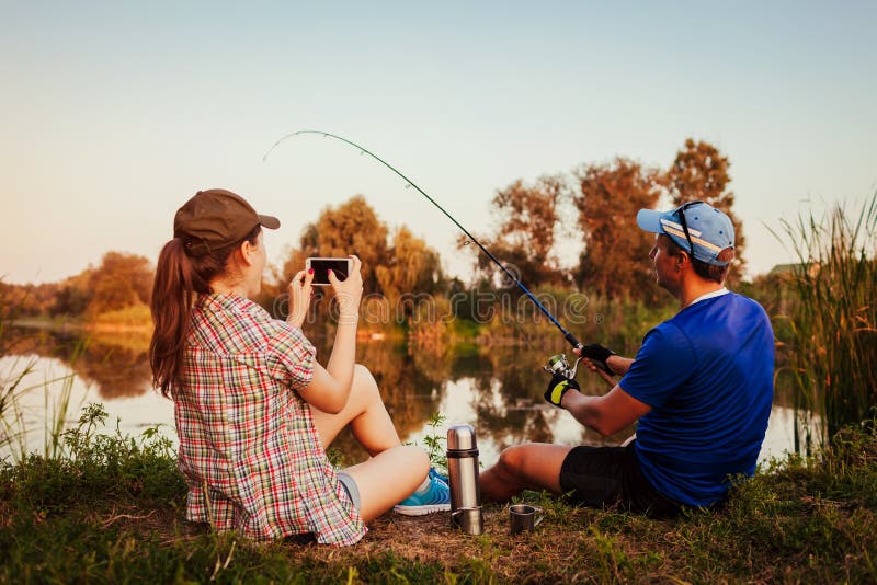 Barnet kopplar ihop fiske och drickate på floden på solnedgången Kvinna som filmar hennes pojkvän som fångar fisken gyckel som ha arkivfoton