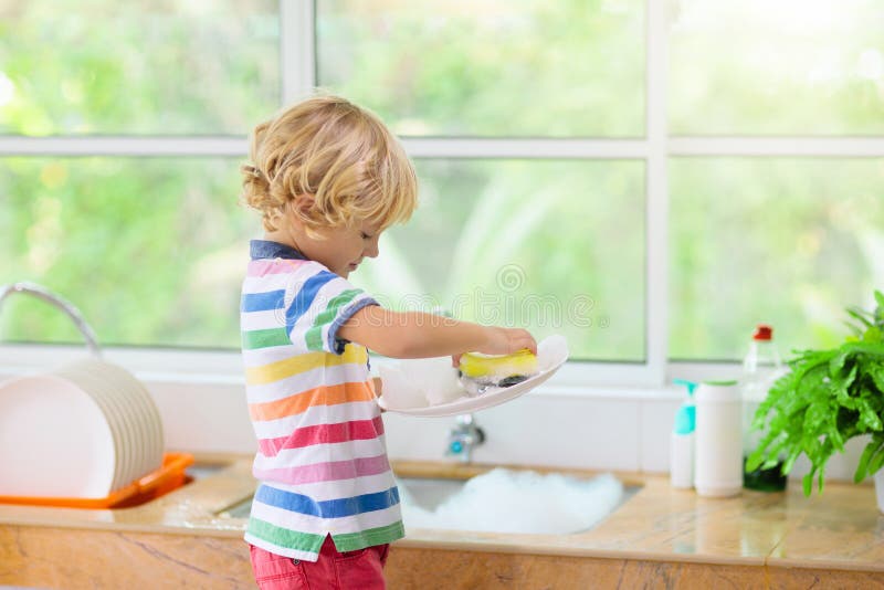 Child washing dishes. Home chores. Kid in white kitchen cleaning plates after lunch at window. Child washing dishes. Home chores. Kid in white kitchen cleaning plates after lunch at window