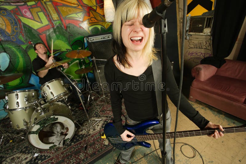 Female bass player screaming into a microphone. Focus on singers face - drummer blurry in the background. Shot with slow shutter speed and strobes - motion blur visible in some areas. Female bass player screaming into a microphone. Focus on singers face - drummer blurry in the background. Shot with slow shutter speed and strobes - motion blur visible in some areas.