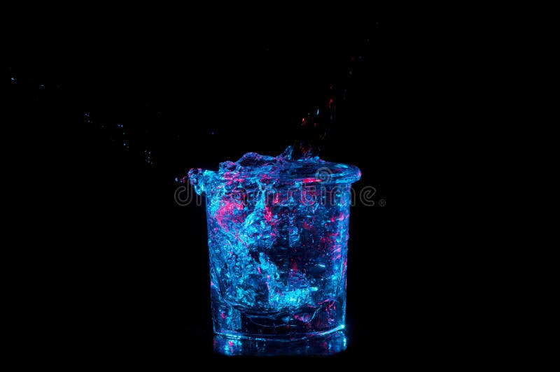 Bascule le débordement en verre avec de l'eau sous les lumières bleues lumineuses avec le rouge sur un fond noir images libres de droits Bascule le débordement en verre avec de l'eau sous les lumières bleues lumineuses avec le rouge sur un fond noir images libres de droits