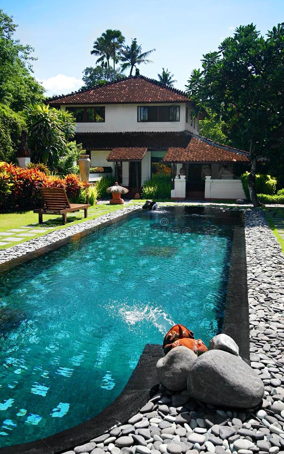 A photograph of a large mansion house with a blue turquoise swimming pool in the front yard, with colorful tropical landscaping in the garden. Taken under sunny blue sky. Vertical color format. A photograph of a large mansion house with a blue turquoise swimming pool in the front yard, with colorful tropical landscaping in the garden. Taken under sunny blue sky. Vertical color format.