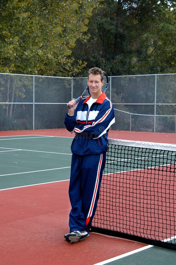 Male teen leans against court net. He is wearing high school varsity uniform and has a racket over his shoulder. Male teen leans against court net. He is wearing high school varsity uniform and has a racket over his shoulder.