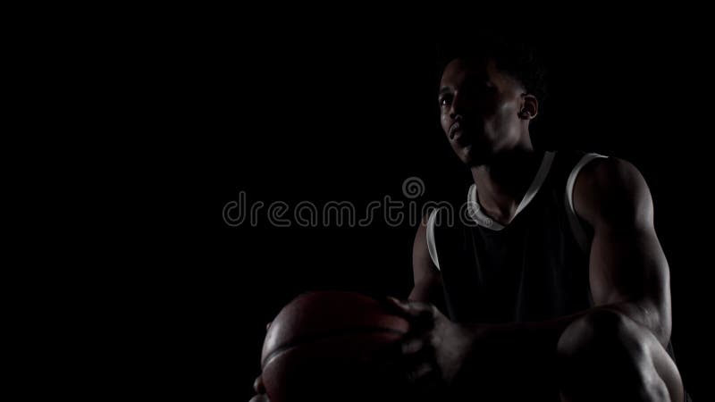Basketbollspelare som sitter på bänk. Framtidsutsikter. Koncentrerad afrikansk amerikansk idrottsman mot svart bakgrund lager videofilmer