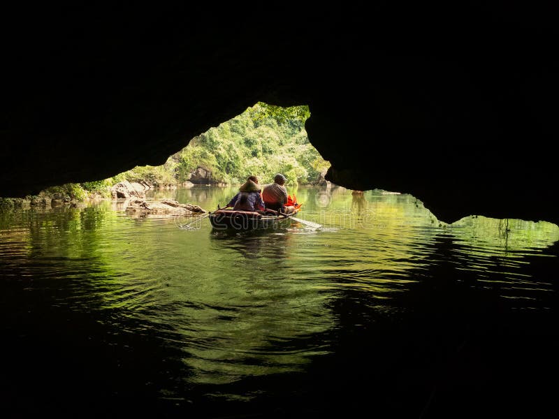 Boat filled with tourists and local guide in bamboo hat exploring caves on a river surrounded by beautiful nature, mountains and jungle. View from behind from a cave, contrast of dark black and bright green light. Boat filled with tourists and local guide in bamboo hat exploring caves on a river surrounded by beautiful nature, mountains and jungle. View from behind from a cave, contrast of dark black and bright green light