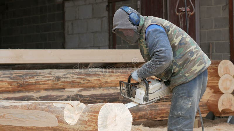 Bauarbeiter schneidet hölzerne Kettensäge für zukünftiges Haus Schutzmaske und Kopfhörer auf dem Kopf des Erbauers und des a