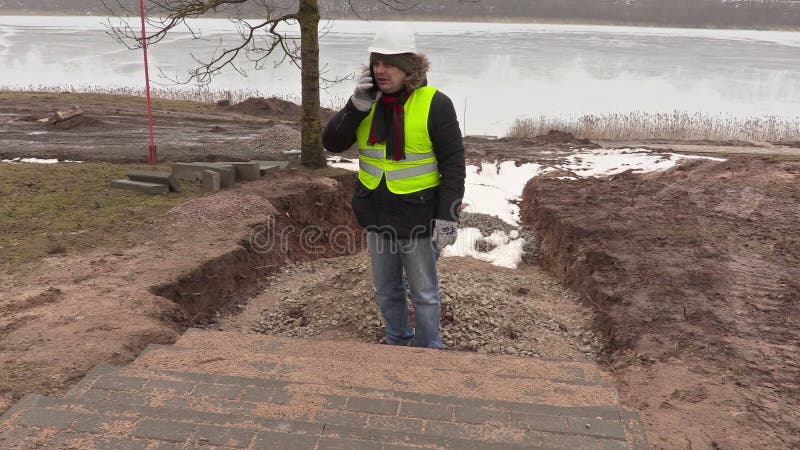 Bauingenieur, der am intelligenten Telefon nahe unfertiger Pflasterung spricht stock footage