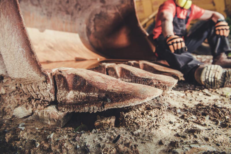 Construction Site Break. Caucasian Worker Taking Short Break and Relax on the Iron Excavator Bucket. Construction Site Break. Caucasian Worker Taking Short Break and Relax on the Iron Excavator Bucket