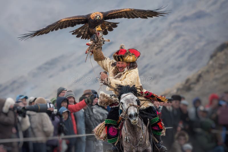 Bayan-Ulgii, Mongolia - October 01, 2017: Golden Eagle Festival. Triumphant Mongolian Hunter Berkutchi In Traditional Clothes Riding On Horse, Holding A Golden Eagle With Prey On Its Hand. Triumph Of Victory. Bayan-Ulgii, Mongolia - October 01, 2017: Golden Eagle Festival. Triumphant Mongolian Hunter Berkutchi In Traditional Clothes Riding On Horse, Holding A Golden Eagle With Prey On Its Hand. Triumph Of Victory