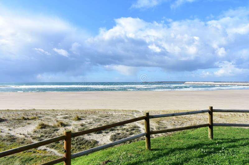 Beach with grass, wooden fence and industrial port. Sea with waves and cloudy sky, Sabon, Spain.