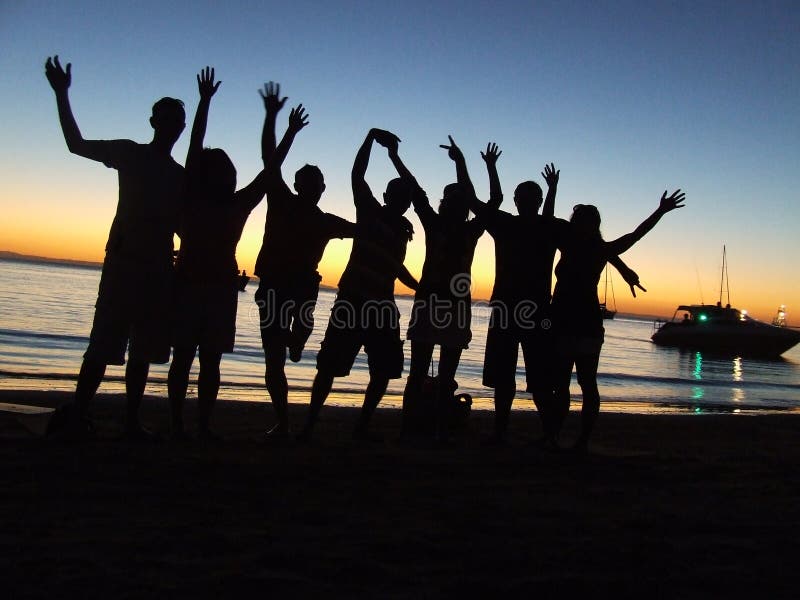 Silhouette di un gruppo di gente allegra una festa su una spiaggia al tramonto, un yacht in background.