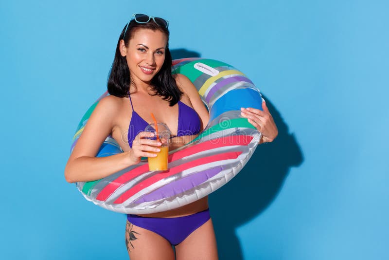 Beach style. Woman in bikini wearing swim ring and sunglasses up the head standing isolated on blue wall with smoothie royalty free stock photos Beach style. Woman in bikini wearing swim ring and sunglasses up the head standing isolated on blue wall with smoothie royalty free stock photos