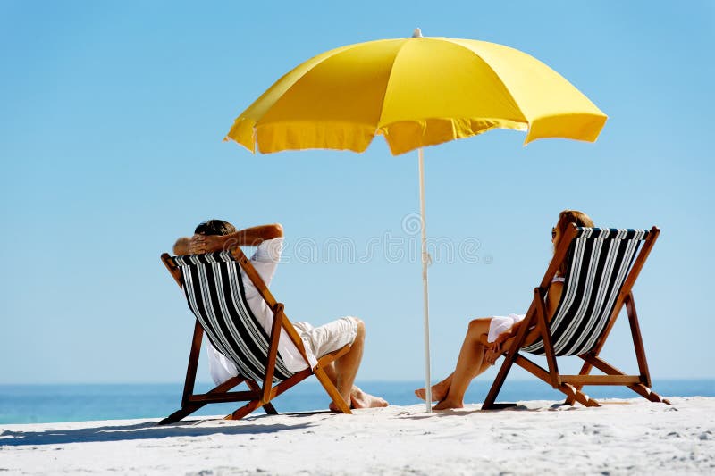 Spiaggia estate coppia in vacanza sull'isola di vacanza per rilassarsi al sole sulle loro sedie a sdraio sotto un ombrello giallo.