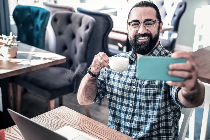 Beaming bearded man feeling relaxed making selfie and drinking coffee stock photos Beaming bearded man feeling relaxed making selfie and drinking coffee stock photos