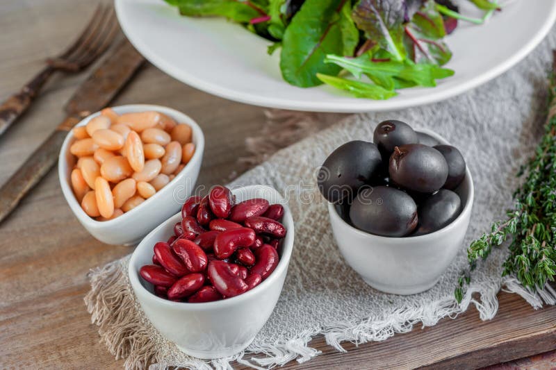 Beans are white and red with large black olives in white small cups with green leaves of arugula and Lollo Ross prepared. For salad with thyme. Prevention of stock image Beans are white and red with large black olives in white small cups with green leaves of arugula and Lollo Ross prepared. For salad with thyme. Prevention of stock image