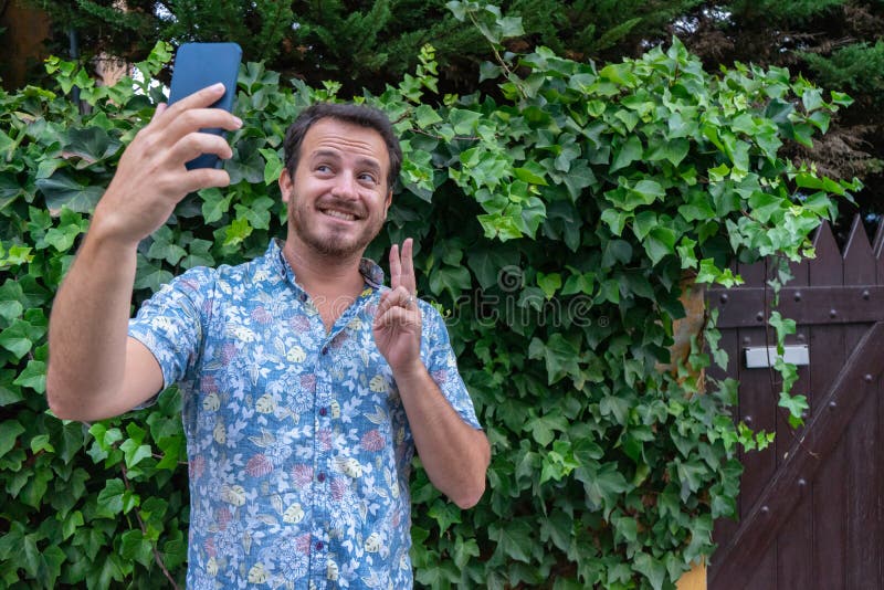 Bearded Boyfriend with modern shirt making a video call. Hipster Guy with beard take a selfie with smart phone and victory fingers