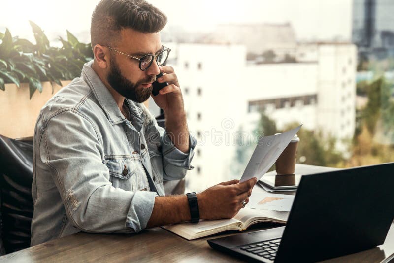 Bearded businessman hipster talking on phone while sitting at desk in office, sad looking at documents. Fall in profits.