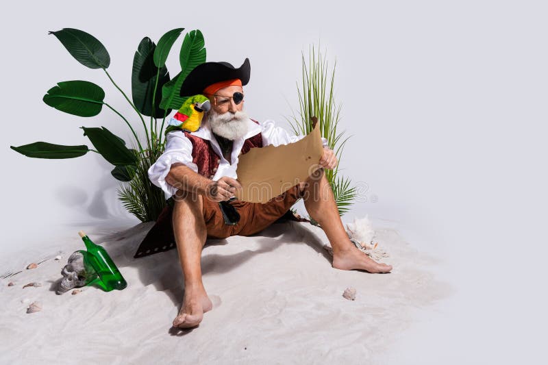 Senior male pirate reading map on beach with lush greenery and bottle stock image