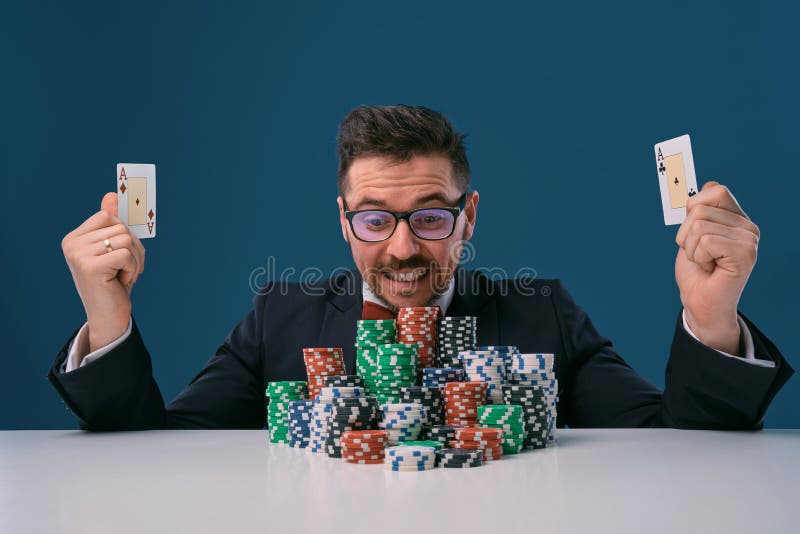 Fellow in glasses, black suit sitting at table with stacks of chips, holding two playing cards, posing on blue royalty free stock photos