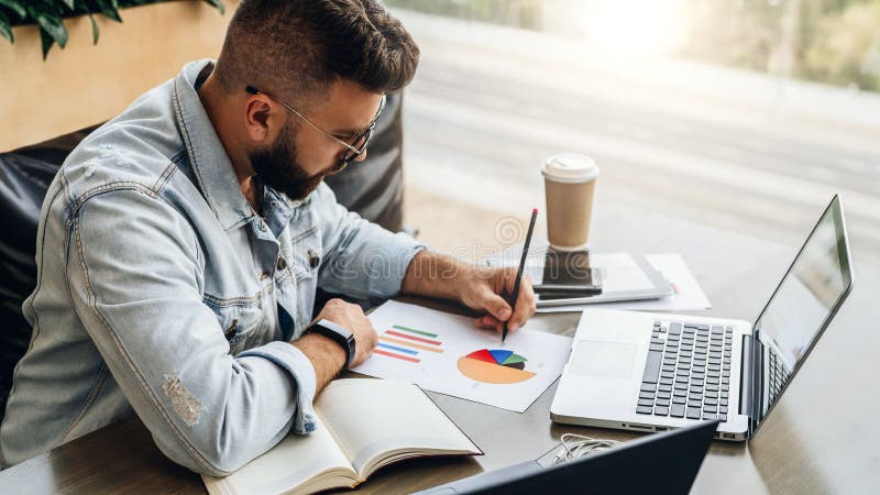 Bearded hipster man sits at table,working on laptop, and makes notes in chart, graph, diagram.Businessman analyzes data. stock photo Bearded hipster man sits at table,working on laptop, and makes notes in chart, graph, diagram.Businessman analyzes data. stock photo