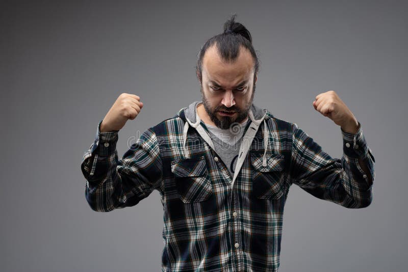 Bearded man with combed hair, standing in checked shirt with clenched fists, as if showing his strength and anger before the fight. Front portrait against grey background. Bearded man with combed hair, standing in checked shirt with clenched fists, as if showing his strength and anger before the fight. Front portrait against grey background