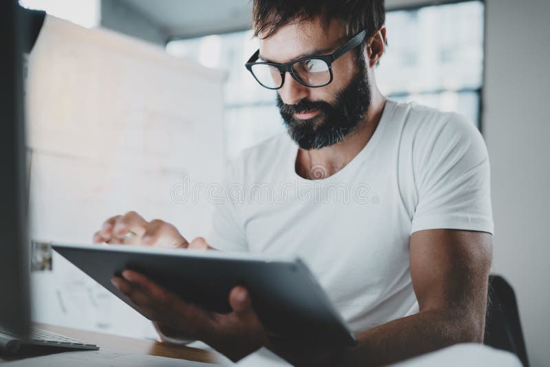 Bearded man in white tshirt working with portable electronic pro tablet computer at modern lightful office.Horizontal