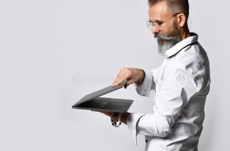 Bearded mature man in a white shirt with a sly smile closes a laptop on a gray background.