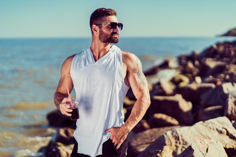 Bearded tattooed manat the resort in a white shirt and sunglasses, sitting on a rock, drink cocktail on the sea stock images Bearded tattooed manat the resort in a white shirt and sunglasses, sitting on a rock, drink cocktail on the sea stock images