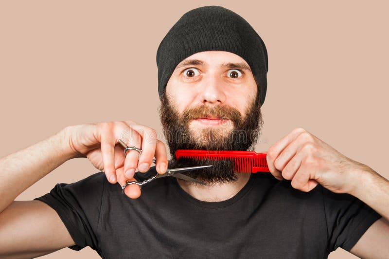 Bearded young guy in hat with comb and scissors cut his beard. Isolated on brown background stock image Bearded young guy in hat with comb and scissors cut his beard. Isolated on brown background stock image