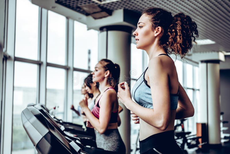 Beau groupe d'amis de jeunes femmes s'exerçant sur un tapis roulant au gymnase moderne lumineux images stock Beau groupe d'amis de jeunes femmes s'exerçant sur un tapis roulant au gymnase moderne lumineux images stock