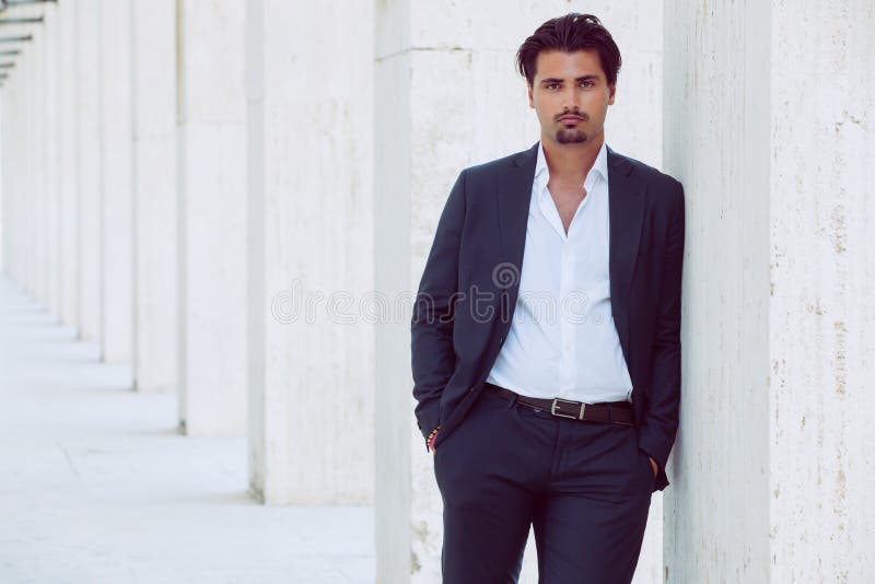 Beau jeune homme élégant extérieur pose un penchement sur une colonne le long d'un corridor. Cheveux à la mode. images stock