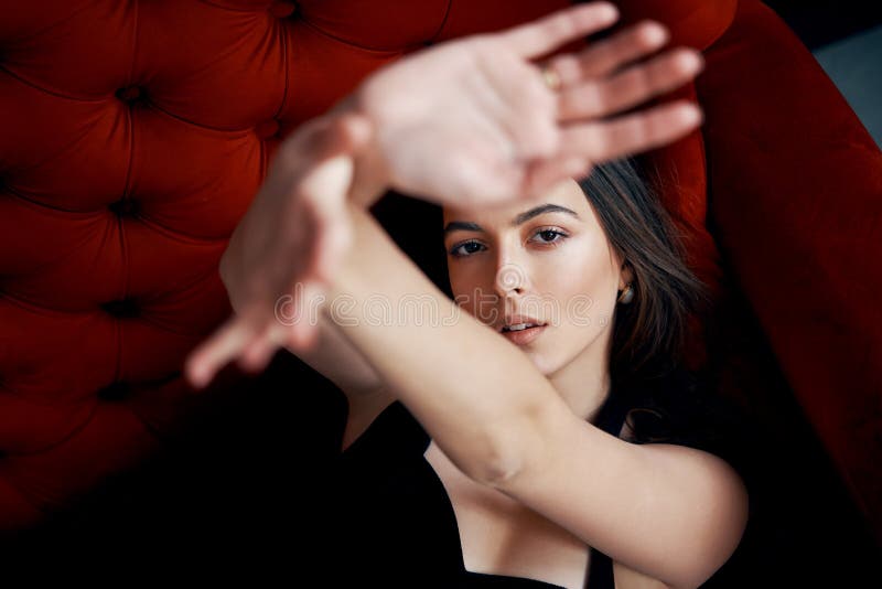 Beau portrait sensuel de femme regardant par un cadre de mains posant dans la robe noire sexy sur le sofa rouge photo libre de droits Beau portrait sensuel de femme regardant par un cadre de mains posant dans la robe noire sexy sur le sofa rouge photo libre de droits
