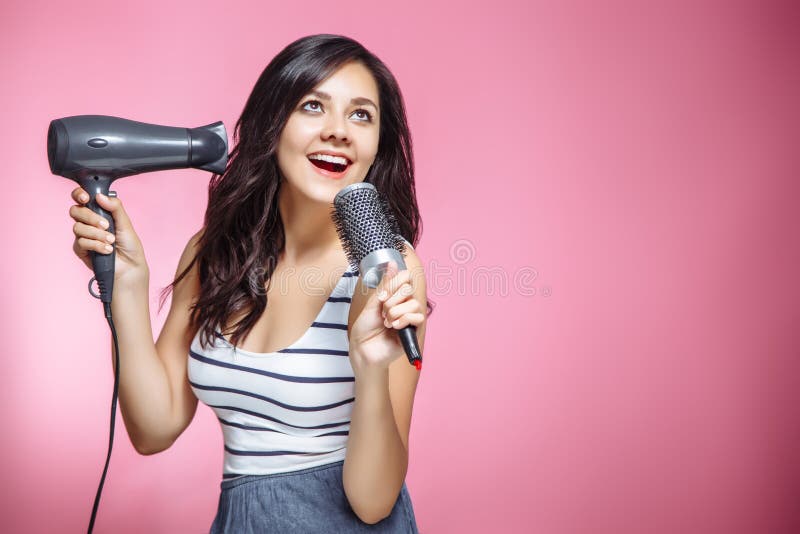 Beau sentiment de jeune femme heureux et chantant tout en employant un hairdryer et une brosse à cheveux sur le fond rose photo stock Beau sentiment de jeune femme heureux et chantant tout en employant un hairdryer et une brosse à cheveux sur le fond rose photo stock