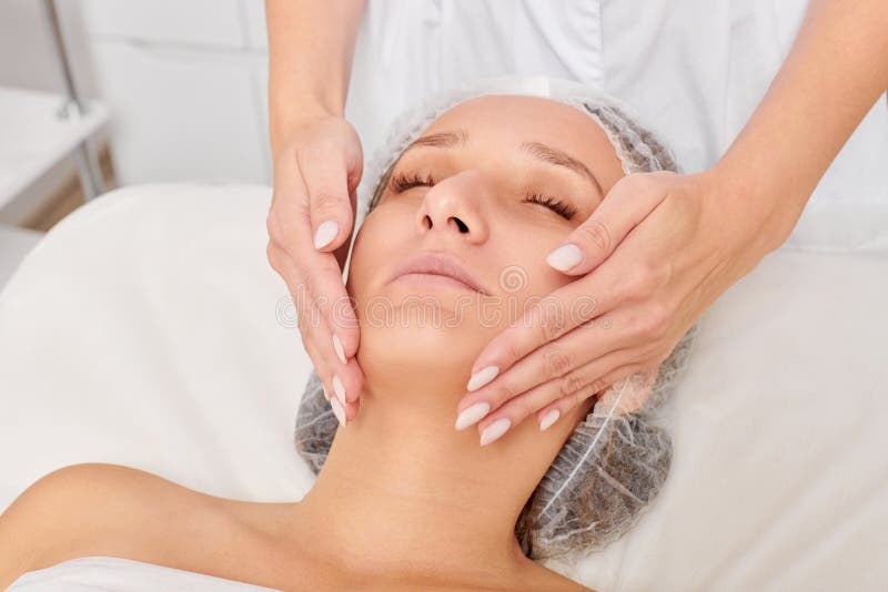 Beautician Massages Cream Mask Into Woman Face Skin For Rejuvenation Procedure In Beauty Salon