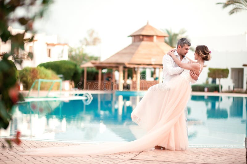 Newly married couple after wedding in luxury resort. Romantic bride and groom relaxing near swimming pool. Honeymoon. Tropical country. Newly married couple after wedding in luxury resort. Romantic bride and groom relaxing near swimming pool. Honeymoon. Tropical country.