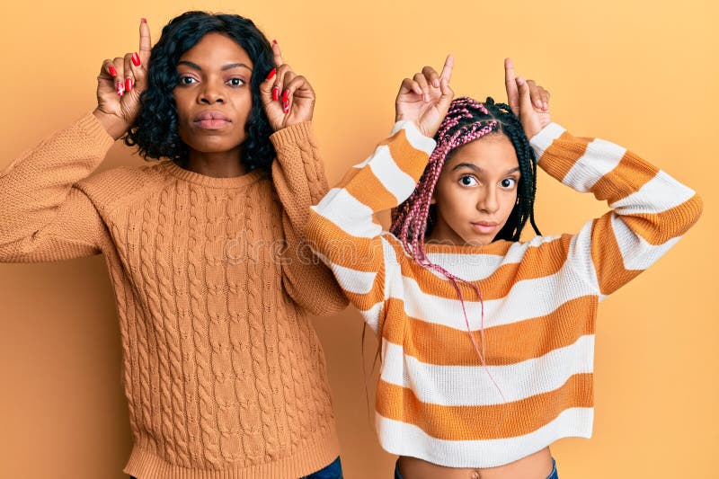 Beautiful african american mother and daughter wearing wool winter sweater doing funny gesture with finger over head as bull horns