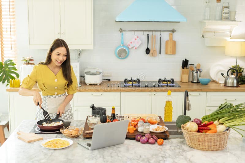 A beautiful Asian girl makes breakfast by looking at how to make it with her laptop computer with a happy face. At her own house