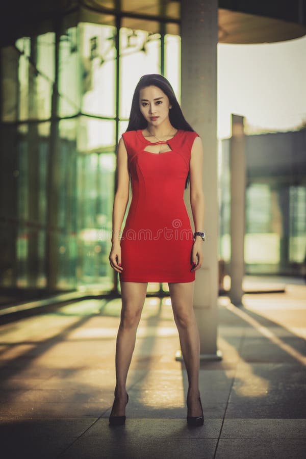 Beautiful Asian girl model in red dress posing at the modern glass style city background. Sunny day.
