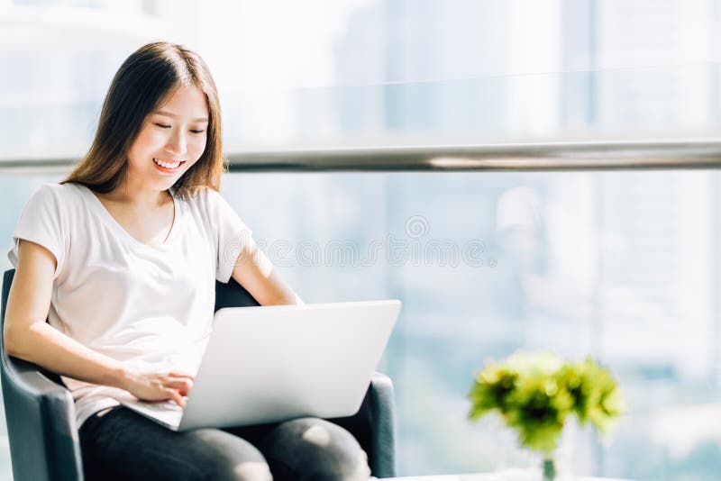 Beautiful Asian girl using laptop computer, smiling happily. College student or freelance worker in modern office. Education or technology or startup business concept with copy space. Beautiful Asian girl using laptop computer, smiling happily. College student or freelance worker in modern office. Education or technology or startup business concept with copy space