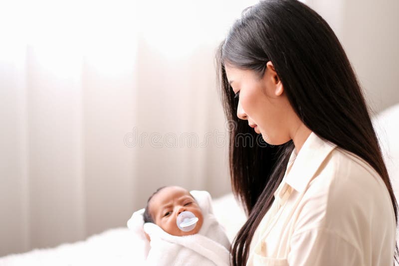 Beautiful Asian mother is holding and look at her newborn baby with love in front of white curtain with day light stock photo Beautiful Asian mother is holding and look at her newborn baby with love in front of white curtain with day light stock photo