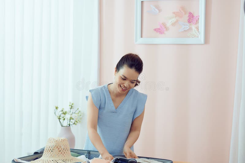 Beautiful asian woman packing summer clothes in suitcase smiling looking to the side and staring away thinking