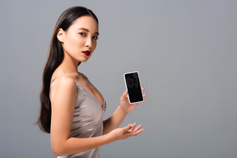 Asian Woman In Satin Dress With Red Lips Holding Smartphone With Blank Screen On Grey Stock