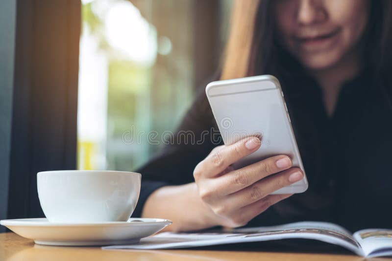 A beautiful Asian woman with smiley face holding and using smart phone while reading magazines with coffee cup on wooden table stock photography A beautiful Asian woman with smiley face holding and using smart phone while reading magazines with coffee cup on wooden table stock photography
