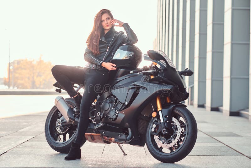 A beautiful biker girl leaning on her superbike outside a building.