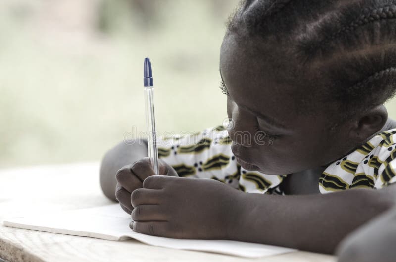 Cute African schoolgirl doing her homework at school: beautiful black girl writing and learning activity with a blue pen. She`s sitting in her desk doing her calculating work in her exercise book.