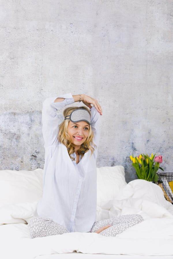 Beautiful blond and young woman, wearing sleeping mask, sitting in bed and doing yoga exercises in the morning royalty free stock image