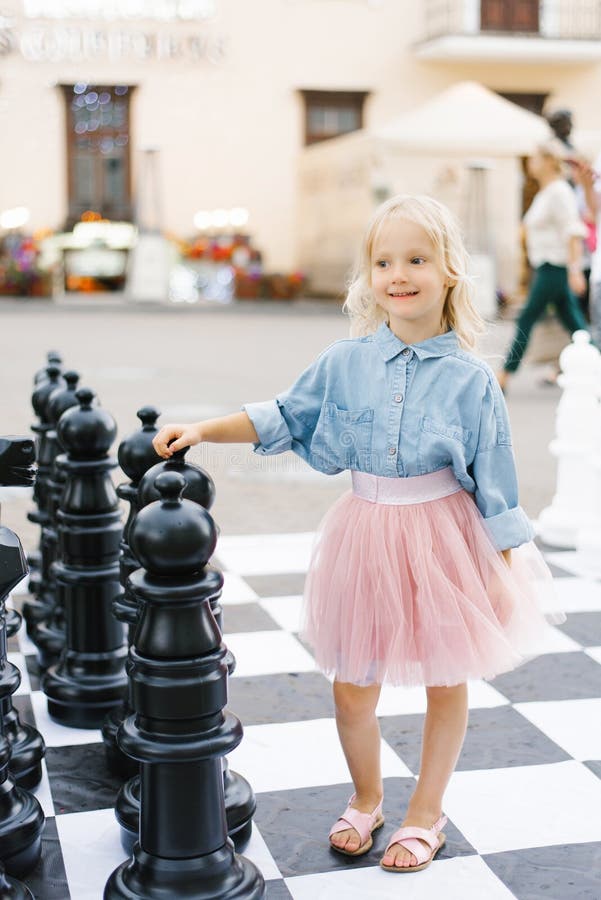 A beautiful blonde girl of five is standing next to a large chess set in the Park